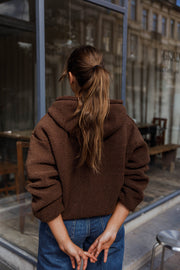 Veste polaire zippée à capuche marron portée par une femme, vue de dos, capuche visible, mains jointes dans le dos, queue de cheval