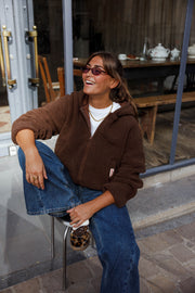 Veste polaire zippée à capuche marron portée par une femme, jambe levée contre la vitrine, éclat de rire, lunettes de soleil, jean large