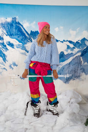 Pull torsadé légèrement loose porté par une femme, silhouette entière, pantalon de ski rose, bâtons