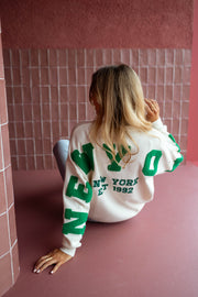 Pull loose à logo dos écru porté par une femme, assise, mur rose, lettres vertes