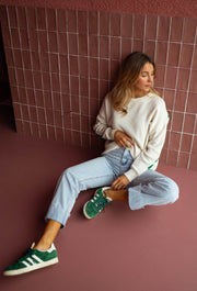 Pull loose à logo dos écru porté par une femme, assise, mur rose, jean