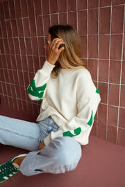Pull loose à logo dos écru porté par une femme, assise, mur rose, profil