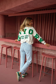 Pull loose à logo dos écru porté par une femme, vue de dos, tabourets roses, bar