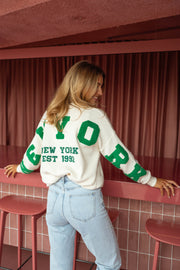 Pull loose à logo dos écru porté par une femme, vue de dos, lettres vertes, tabourets roses