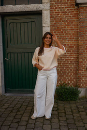 Pull loose à manches courtes porté par une femme, silhouette entière, pantalon blanc, adossée à une porte verte