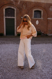 Pull loose à manches courtes porté par une femme, silhouette entière, pantalon blanc, devant un mur en briques