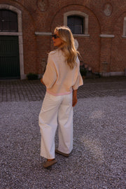 Pull loose à manches courtes porté par une femme, vue de dos, pantalon blanc, devant un mur en briques