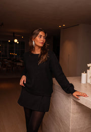 Pull loose à dos ouvert noir porté par une femme, vue de face, short noir, comptoir