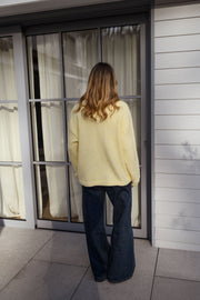 Pull col rond légèrement ample porté par une femme, vue de dos, rideaux blancs, jean foncé