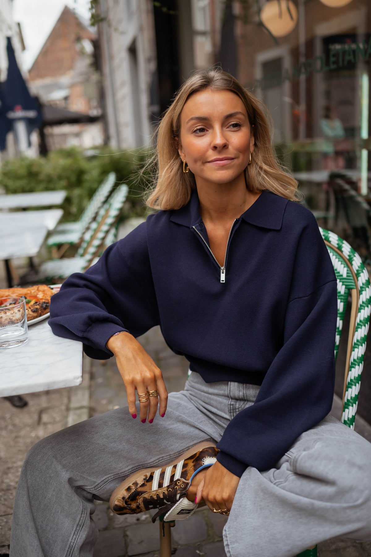 Pull court à col zippé bleu porté par une femme, assise, terrasse, chaises vertes