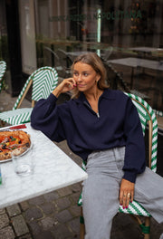 Pull court à col zippé bleu porté par une femme, assise, table, pizza