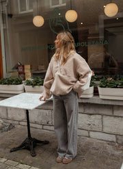 Pull ample à capuche zippé beige porté par une femme, vue de trois-quarts, table, pantalon gris