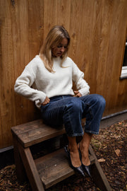 Pull à dos ouvert fleuri écru porté par une femme, assise, tabouret en bois, bottines