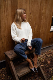 Pull à dos ouvert fleuri écru porté par une femme, assise, mur en bois, jean bleu