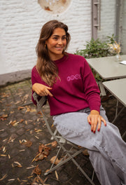 Pull à logo au dos bordeaux porté par une femme, assise, chaise de jardin, jean gris