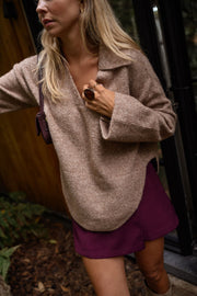 Pull doux col V loose beige porté par une femme, vue de trois-quarts, mur en bois, main sur le col