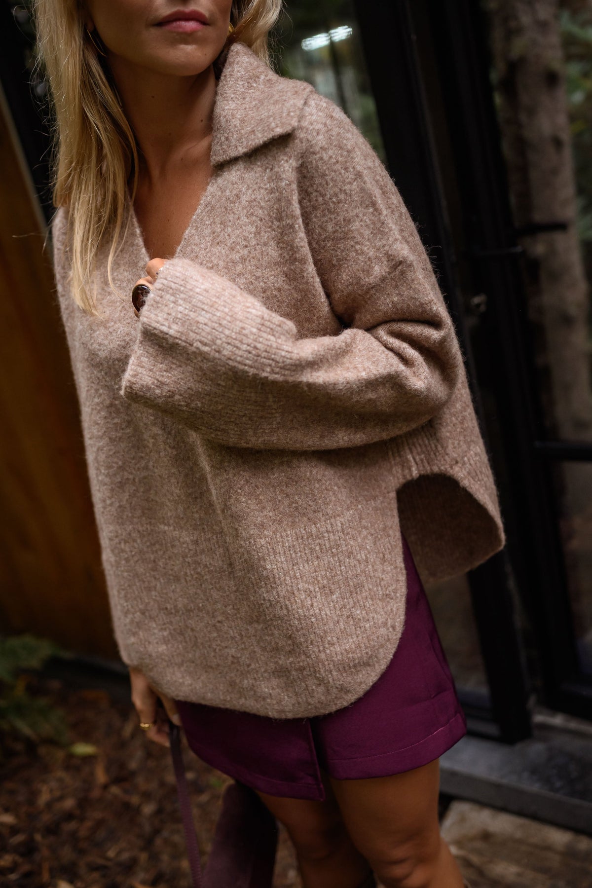 Pull doux col V loose beige porté par une femme, vue de face, mur en bois, jupe bordeaux