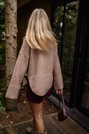 Pull doux col V loose beige porté par une femme, vue de dos, véranda, cheveux blonds