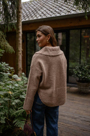 Pull doux col V loose beige porté par une femme, vue de dos, cabane, jean bleu
