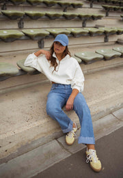 Pull court à cordons écru porté par une femme, assise, gradins, casquette bleue