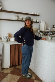 Pull doux à boutons latéraux bleu marine porté par une femme, silhouette entière, près d'un comptoir