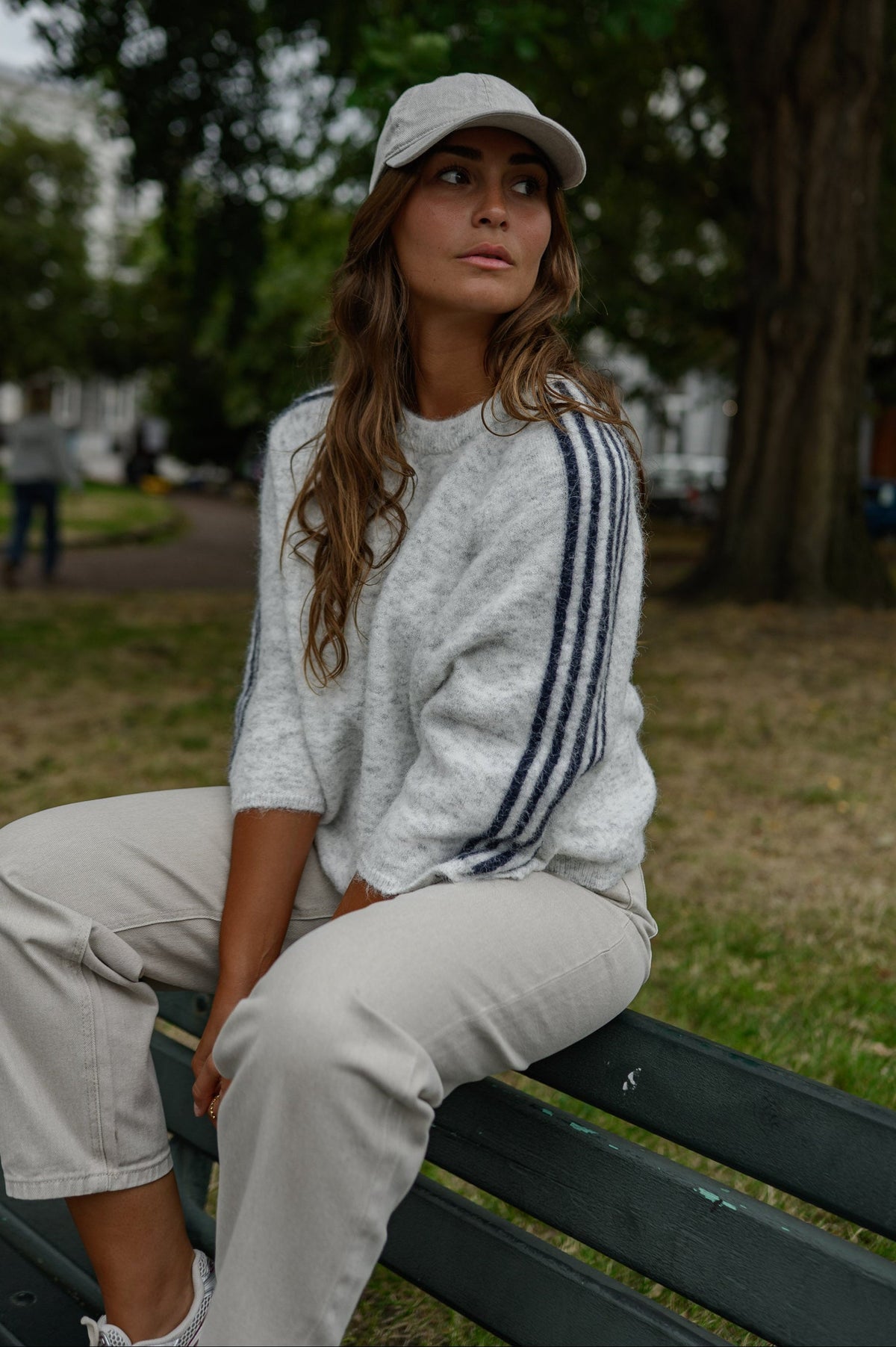 Pull à bandes manches trois-quarts porté par une femme, assise, sur un banc, casquette grise, pantalon clair