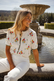 Pull fleuri à manches courtes porté par une femme, assise, margelle de fontaine, pantalon blanc