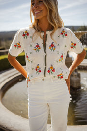 Pull fleuri à manches courtes porté par une femme, vue de face, mains sur les hanches, pantalon blanc