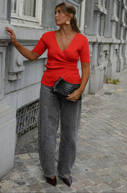 Pull cache-cœur manches courtes porté par une femme, silhouette entière, jean gris, sac noir, adossée à un mur gris
