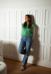Pull sans manches doux vert porté par une femme, vue de face, appuyée au mur, jean