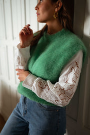 Pull sans manches doux vert porté par une femme, vue rapprochée, manches en dentelle, canapé