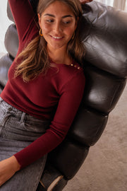 Pull moulant à boutons d’épaule bordeaux porté par une femme, assise, fauteuil, sourire