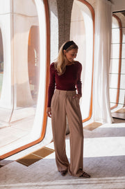 Pull moulant à boutons d’épaule bordeaux porté par une femme, silhouette entière, baie vitrée, pantalon beige