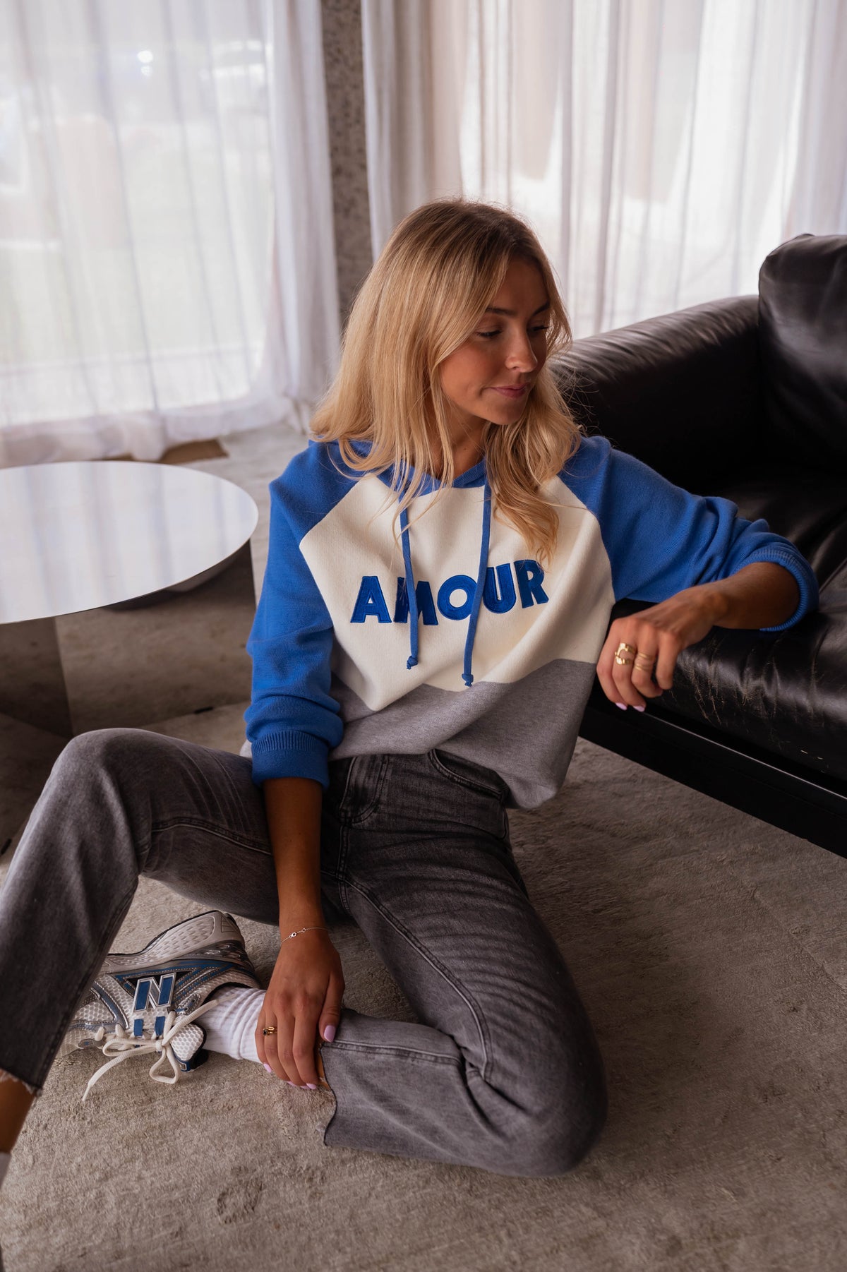 Pull doux à capuche bleu porté par une femme, assise, canapé noir, jean gris