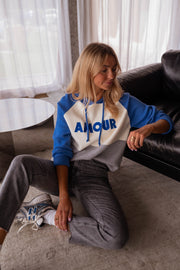 Pull doux à capuche bleu porté par une femme, assise, canapé noir, jean gris