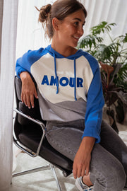 Pull doux à capuche bleu porté par une femme, assise, chaise noire, sourire