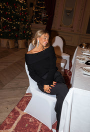 Pull loose à col bardot noir porté par une femme, vue de trois-quarts, chaise blanche, table dressée