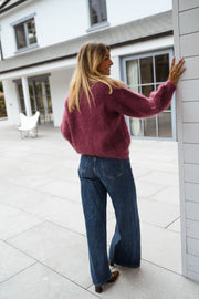 Pull ample aubergine col rond porté par une femme, vue de trois-quarts, mur gris, main sur le mur
