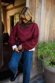 Pull ample doux col rond bordeaux porté par une femme, vue de trois-quarts, porte en bois, mains sur le sac