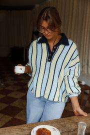 Pull ample à col boutonné bleu clair et crème à rayures porté par une femme, vue de face, main sur la table