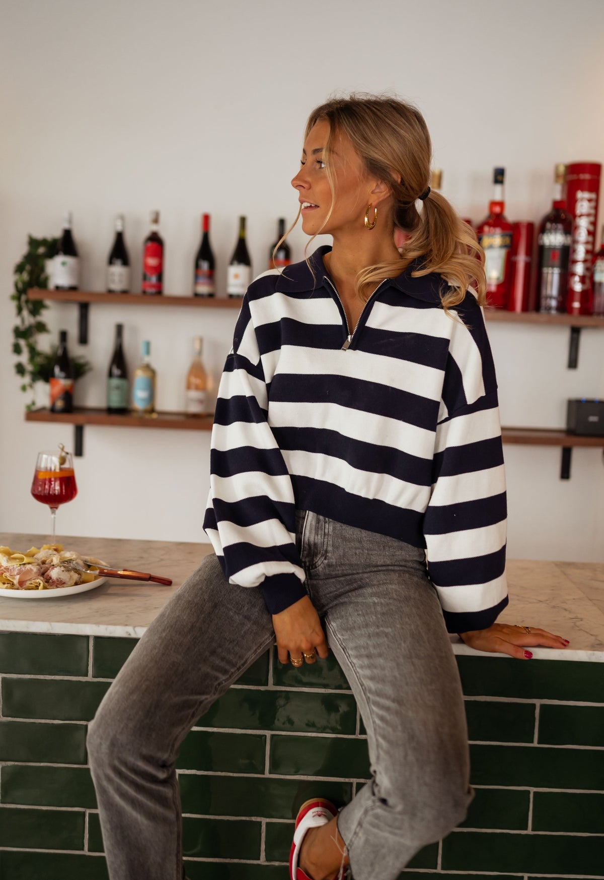 Pull rayé à col zippé porté par une femme, vue de face, bar, étagère de bouteilles
