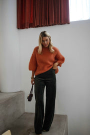 Pull col rond manches trois-quarts orange porté par une femme, vue de face, rideau rouge, jean noir