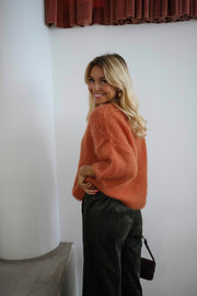 Pull col rond manches trois-quarts orange porté par une femme, vue de trois-quarts, rideau rouge, sac noir