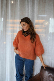 Pull col rond manches trois-quarts orange porté par une femme, vue de face, rideaux blancs, foulard noir