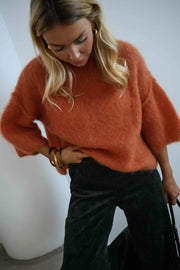 Pull col rond manches trois-quarts orange porté par une femme, vue rapprochée, jean noir, cheveux blonds baissés
