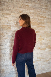 Pull doux légèrement court bordeaux porté par une femme, vue de dos, mur en pierre, jean