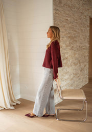 Pull doux légèrement court bordeaux porté par une femme, vue de profil, chaise en métal, pantalon gris