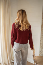 Pull doux légèrement court bordeaux porté par une femme, vue de dos, cheveux blonds, pantalon gris