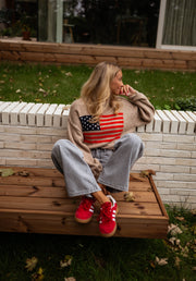 Pull à drapeau col rond camel porté par une femme, assise, banc en bois, baskets rouges