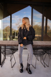 Pull ample zippé à capuche noir porté par une femme, vue de face, bottines noires, table